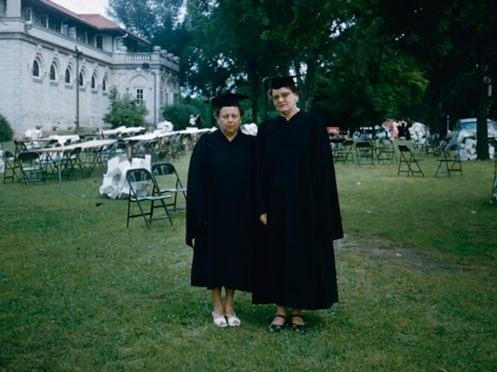 Formatura: duas formandas de beca e capelo no gramado, com as cadeiras da cerimônia atrás
